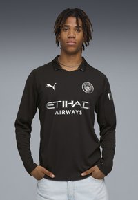 Un tânăr cu dreadlocks purtând un tricou cu mânecă lungă negru de la Manchester City, cu logo-ul Etihad Airways, stând cu mâinile în buzunarele pantalonilor din blugi deschiși la culoare.