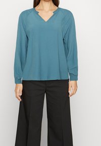 Blouse turquoise à manches longues avec un décolleté en V et des détails de col volanté, associée à un pantalon noir à jambes larges ; texture de tissu lisse.