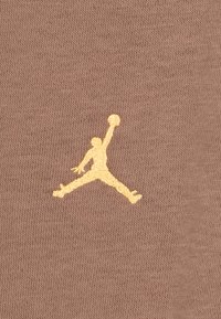 Ruda audinio medžiaga su iškilniu auksiniu Jumpman logotipu, vaizduojančiu dinamišką pozą su krepšiniu, ant tekstūruoto paviršiaus.