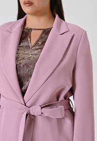 Lichtroze blazer met een riemdetail, gestructureerde revers en gladde stof. Daaronder een glanzend bruine top met een keyhole-halslijn.