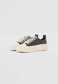 Canvas-Sneaker mit einem dunkelgrauen Obermaterial, einer kremfarbenen Gummikappe, dicker Sohle und beigen Schnürsenkeln. Schlupfdesign mit einer Zuglasche an der Rückseite.
