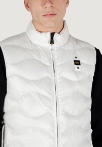 Hvid quilted vest med høj krave, lynlås foran og sorte accenter. Bemærkelsesværdig logo detalje på øverste venstre bryst. Glat tekstur.