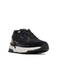 Zapatillas deportivas negras que combinan gamuza y cuero, con detalles texturizados, suela blanca gruesa y un refuerzo en el talón de color marrón en contraste.