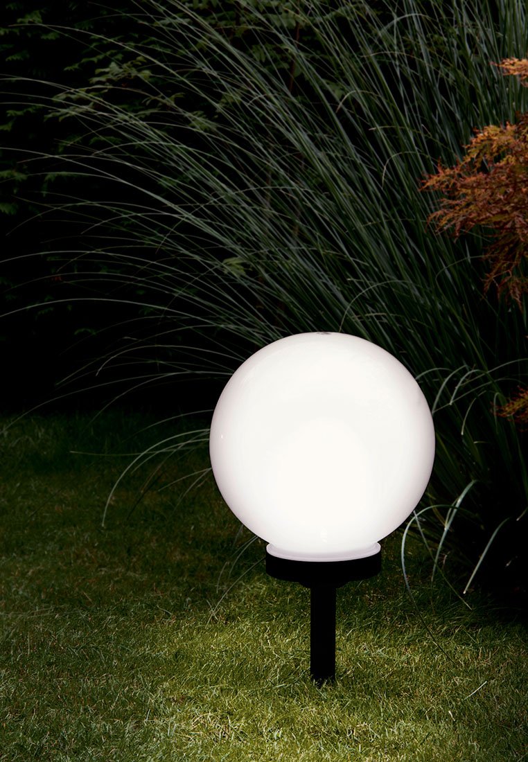 Weiße sphärische Gartenleuchte auf einem schwarzen Pfosten, auf Gras platziert, mit hohen grünen Pflanzen im Hintergrund. Glatte Oberfläche, runde Form.