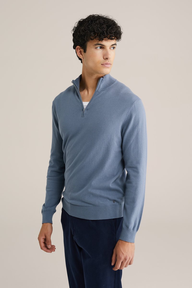 Kék kötött pulóver fél cipzárral, szűkített fazonnal, ribbelt mandzsetta és szegéllyel, puha anyagból készült, az oldalon egy kis logós részlettel.