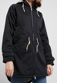 Veste noire à capuche avec fermeture éclair à l'avant, cordons de serrage et accents en cuir. Comprend des poches sur les côtés et une doublure intérieure texturée.