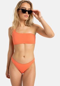Top de bikini orange à une épaule avec un design texturé ; associé à un bas de bikini taille basse assorti. Accessoire de lunettes noires.