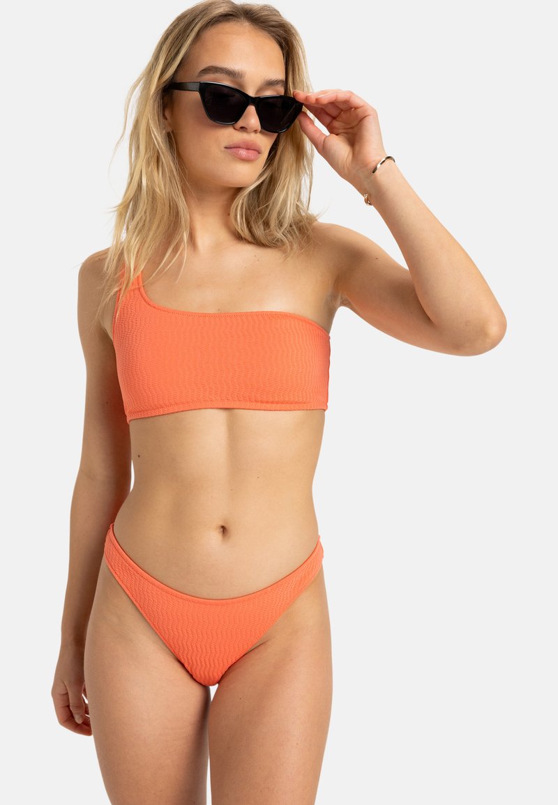 Top de bikini orange à une épaule avec un design texturé ; associé à un bas de bikini taille basse assorti. Accessoire de lunettes noires.
