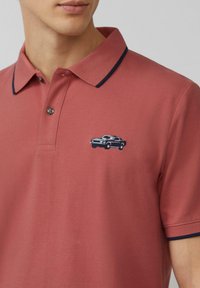 Polo rossa con rifinitura blu scuro di colletto, caratterizzata da un piccolo design di un'auto ricamato in nero sul lato sinistro del petto. Realizzata in tessuto testurizzato.