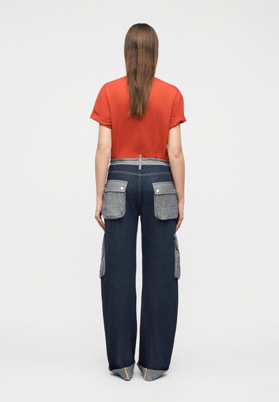 Γυναίκα που στέκεται με την πλάτη, φοράει κόκκινο κοντομάνικο πουκάμισο και σκούρο τζιν με φαρδιά πόδια και μεγάλες γκρι τσέπες στο πίσω μέρος και στα πλευρά.