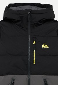 Fekete és szürke párnázott kabát kapucnival, középen fekete cipzárral, a mellzsebnél sárga cipzárral és sárga Quiksilver logóval.