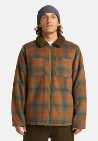 Ruitjes flanellen jas in oranje, blauw en groene tinten met een bruine sherpa kraag en twee borstzakken. Volledig met rits te sluiten.