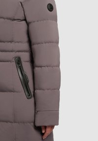 Pufferjakke i mauve med kviltede sektioner, der har en sort lynlåslomme på ærmet og en rund logo-patch på skulderen.