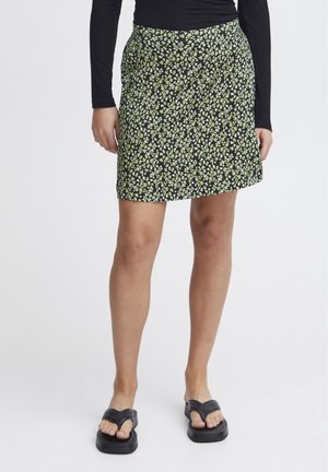 Kvinde iført sort top med lange ærmer, kort sort nederdel med lille gul og hvid blomstret print og sorte platformssandaler.