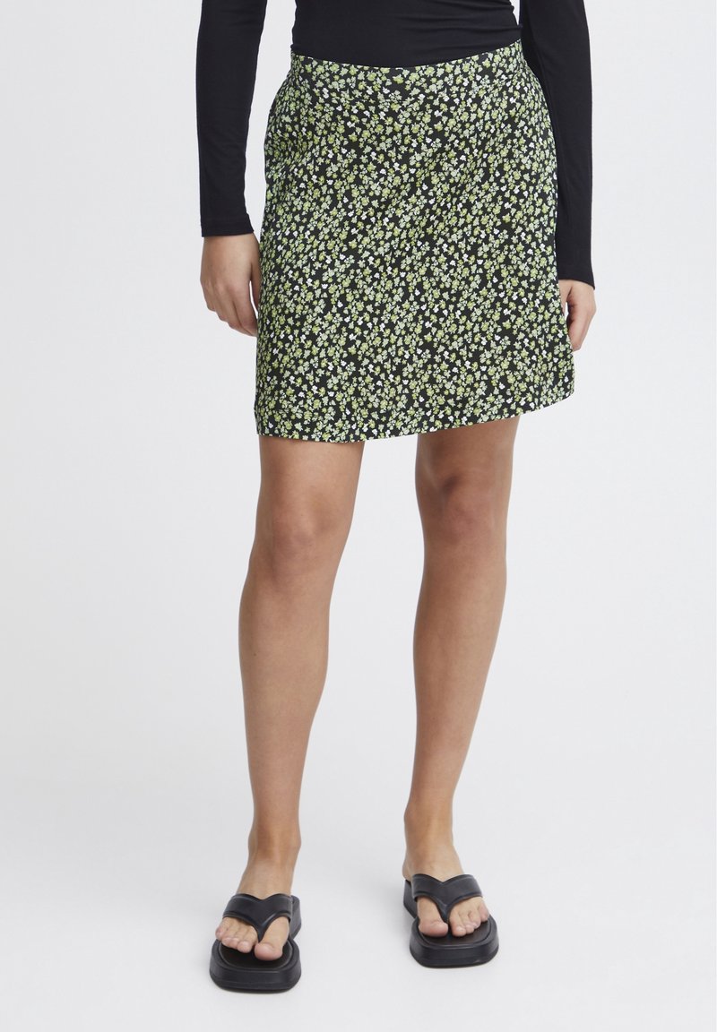 Femme portant un haut noir à manches longues, une jupe courte noire avec un petit imprimé floral jaune et blanc, et des sandales compensées noires.