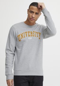 Sudadera gris hecha de una tela suave, con un cuello redondeado y puños acanalados. "UNIVERSIDAD" en letras amarillas-naranjas en negrita a lo largo del pecho.