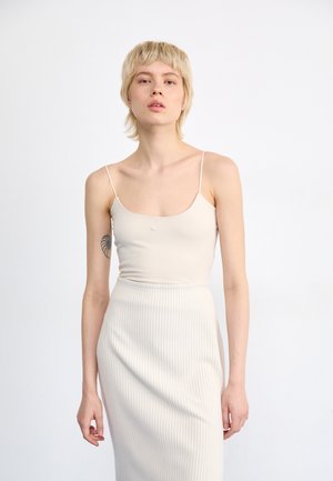 Top sin mangas ajustado en color beige con tirantes finos, que presenta un sutil logo, combinado con una falda midi acanalada del mismo color. Textura de tela suave.