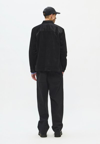 Homme portant une veste en velours côtelé noire, un pantalon noir, une casquette noire et des chaussures sombres, debout de dos sur un fond blanc uni.