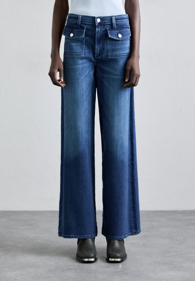 Bredde ben denim jeans i dyb blå, med forreste laplommer, metal knapdetaljer og en glat tekstur. Båret med sorte støvler med hæl.