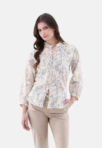 White floral blouse with yellow and gray patterns, button-down front, ruffled neckline, and long puffed sleeves. Paired with beige trousers.