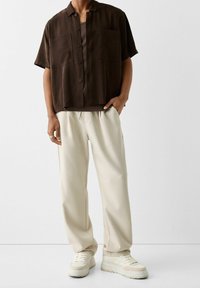 Hnědá krátká košile na knoflíky s dvěma předními kapsami, doplněná krémovými širokými kalhotami a béžovými teniskami.