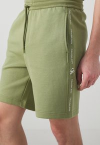 Pantalones cortos de mezcla de algodón verde con cintura elástica y cordón de ajuste, que presentan una marca blanca a lo largo del costado y bolsillos laterales.