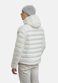 Uomo che indossa un giubbotto imbottito bianco con cappuccio, pantaloni chiari e un berretto grigio a maglia, rivolto di lato con uno sfondo neutro.