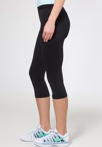 Schwarze Capri-Leggings aus dehnbarem Material mit figurbetonter Passform. Verfügt über einen breiten Bund und kontrastierende Nähte. Getragen mit weißen und türkisfarbenen Sportschuhen.