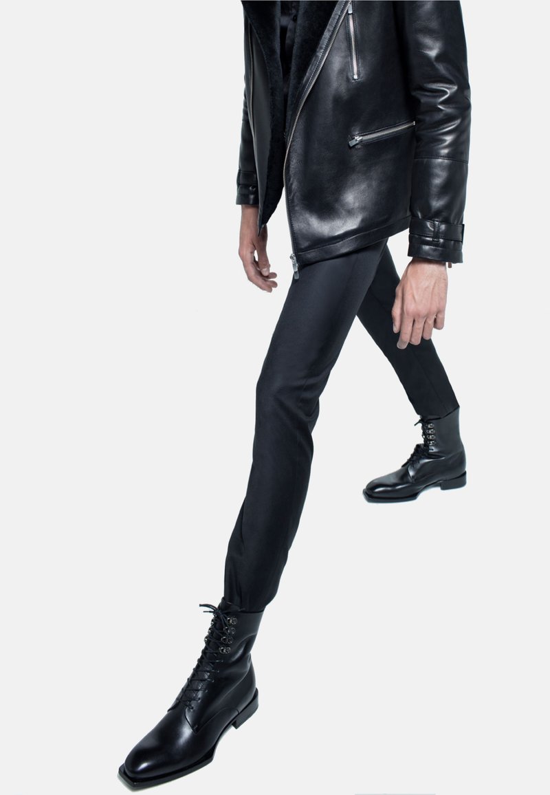 Schwarze Lederjacke mit silbernen Reißverschlüssen, enganliegende schwarze Hose und glänzende schwarze Schnürstiefel mit quadratischer Zehenform.