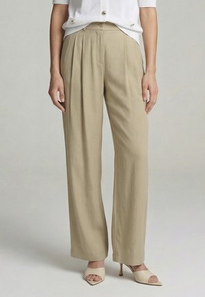 Woman wearing beige wide-leg pants, white buttoned top, and beige open-toe heeled mules, standing in neutral setting.