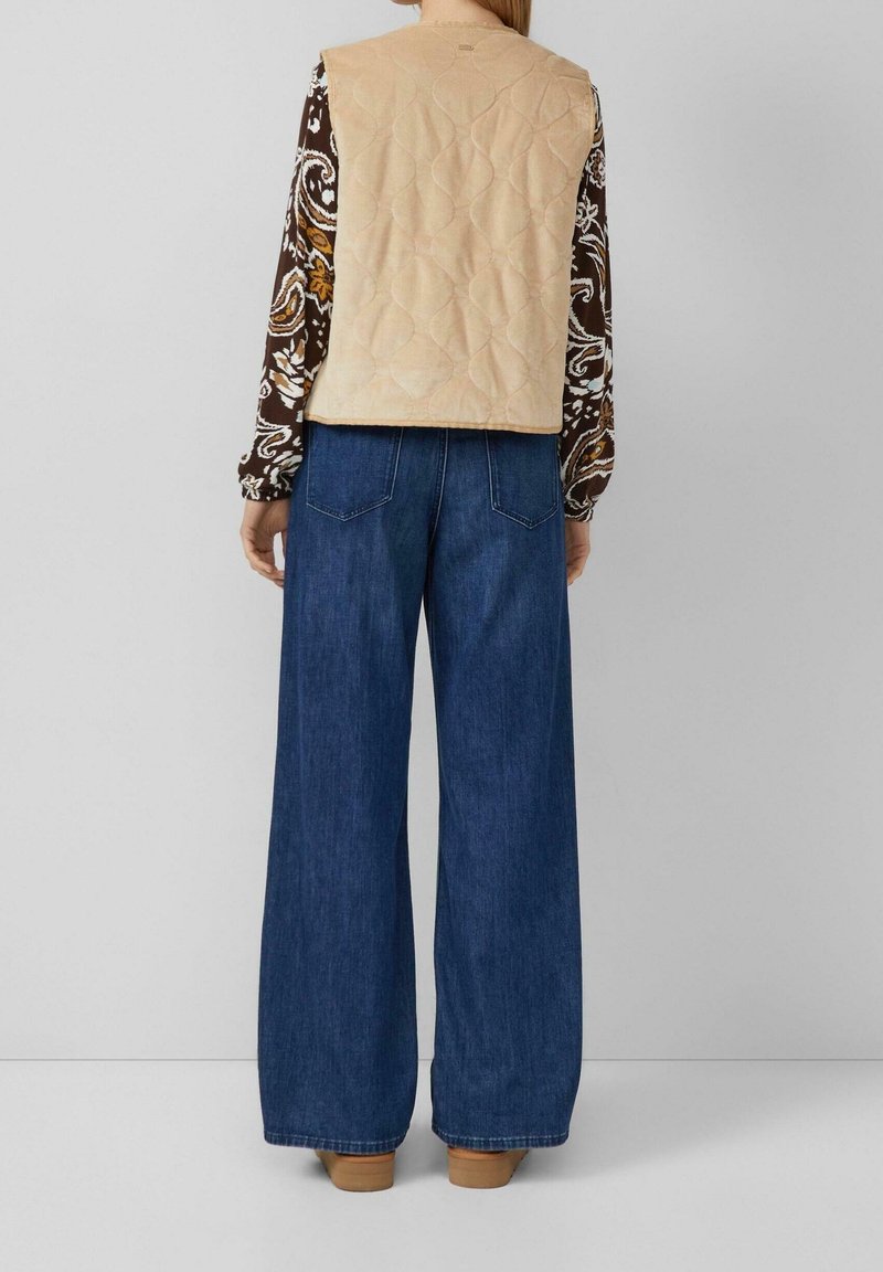 Personne portant un gilet matelassé beige, une chemise à manches longues à motifs, un jean bleu à jambes larges et des chaussures marron, debout devant un fond clair uni.