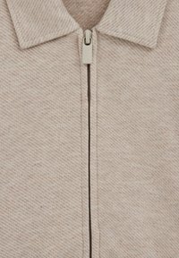 Lysebeige lynlåstrøje med en tekstureret fiskebenmønster og en lille krave. Har en glat lynlås på fronten.