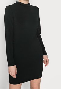 Mustassa pitkähihaisessa mekossa on korkea kaulus, se on valmistettu sileästä kankaasta, ja siinä on tiukka siluetti sekä epäsymmetrinen helma.