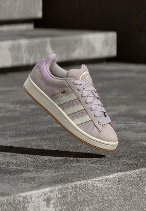 Lavendelfarget Adidas Campus-sneaker med hvite striper og gummisåle som svever over betongtrinn i mykt lys.
