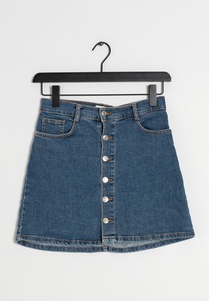 Mini-jupe en jean bleu avec fermeture boutonnée à l'avant, passants pour ceinture et poches, suspendue sur un cintre noir sur fond blanc.