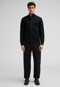 Zwarte zip-up jas met een gestructureerd ontwerp, hoge kraag, ribgebreide manchetten en zoom, gecombineerd met losse zwarte cargobroek en stevige zwarte schoenen.