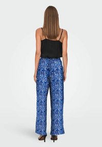 Mujer con cabello castaño liso que lleva una blusa sin mangas negra, pantalones de pierna ancha con patrón azul y blanco, y sandalias de tacón negras, de pie mirando hacia atrás.