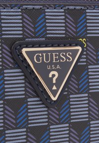 Pung med en sort mønstret overflade og blå og lilla striber. Inkluderer et guld- og sort trekantet logo mærket "GUESS U.S.A."