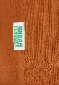 Gros plan sur un tissu côtelé orange avec une étiquette blanche de vêtement indiquant "URBAN CLASSICS" en texte vertical vert.