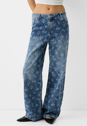 Farkkujen leveälahkeiset housut, joissa on haalistunut kukkakirjailu, yhdistetty mustiin teräväkärkisiin kenkiin ja valkoiseen lyhyeen toppiin. Kuvassa vyötäröstä alaspäin valkoisella taustalla.