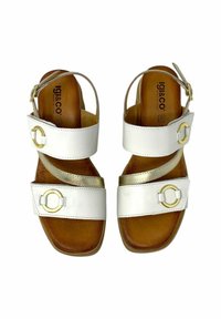Sandali in pelle bianca con accenti dorati, design della fascia curva e suola marrone. Presentano un dettaglio hardware circolare e una cinghia alla caviglia regolabile.