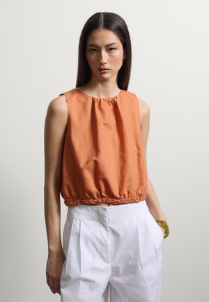 Femme aux cheveux raides et foncés portant un top court orange sans manches avec un ourlet tressé et un pantalon blanc plissé taille haute, debout devant un fond uni.