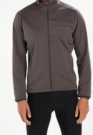 Veste grise à fermeture éclair avec poches sur la poitrine et les côtés portée par une personne, associée à un pantalon noir, sur fond blanc.