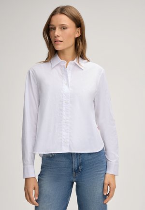 Young woman wearing a plain white button-up long-sleeve shirt and blue jeans, standing against a neutral background.
