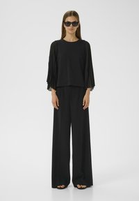 Top nero con maniche lunghe plissettate, abbinato a pantaloni neri a gamba larga. Il tessuto sembra leggero e liscio, con accenti minimi.