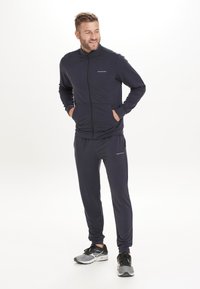 Marinblå träningsoverall i mjukt tyg, med en zipparjacka som har fickor och matchande byxor. Liten logotyp av märket på båda plaggen.