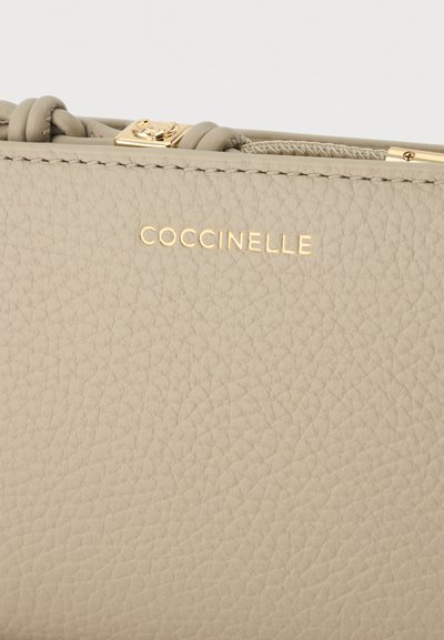 Esquina de bolso de cuero texturizado beige con logo dorado de Coccinelle y una correa de cuero torcida con herrajes dorados.