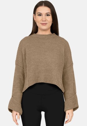 Vrouw met steil bruin haar draagt een losse, geribbelde bruine trui met brede manchetten en een zwarte broek, kijkt recht vooruit tegen een effen achtergrond.