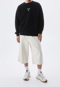 Černá mikina s bílým logem, krémové kraťasy ke kolenům a šedobílé tenisky s hnědými akcenty. Hladké textury a volný střih.