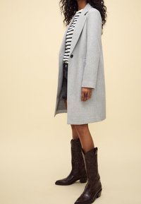 Gray wool coat with a structured design, featuring notched lapels and a single button. Paired with a striped shirt and denim shorts.